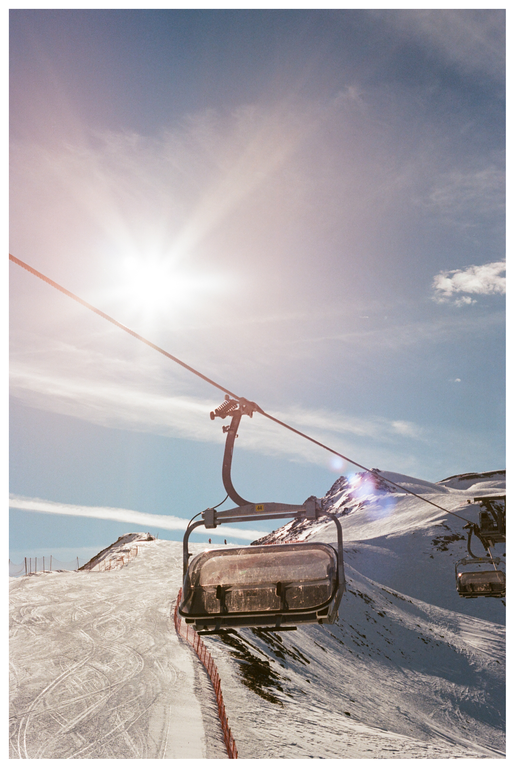 A ski lift chair is visible against a bright sun and a snowy mountain landscape.