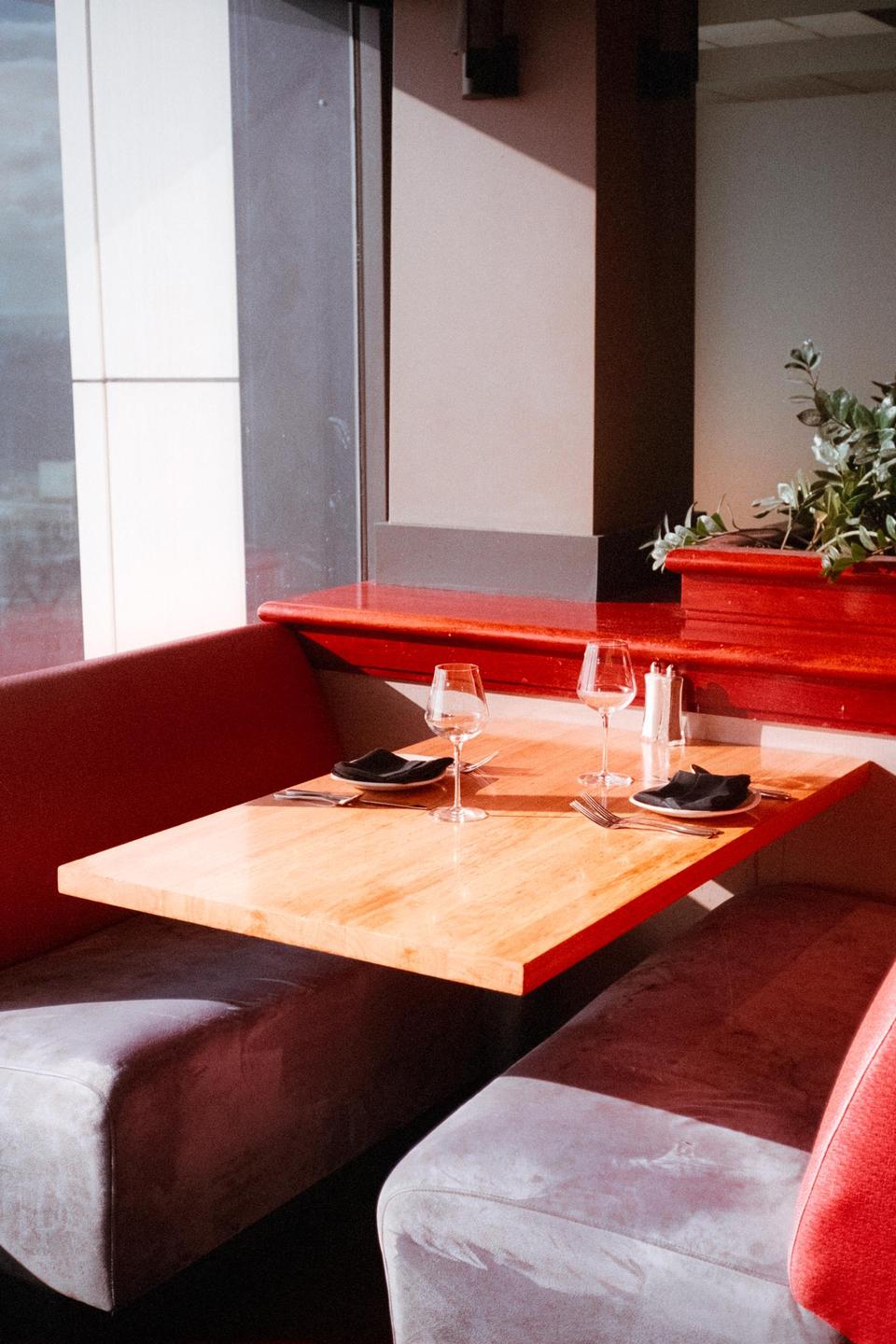 A restaurant booth with a wooden table set for two is illuminated by sunlight streaming through a large window.