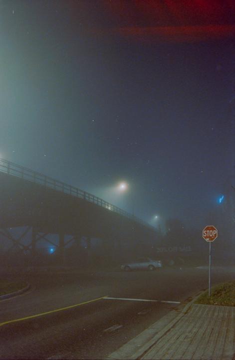 A foggy night scene shows a bridge over a street with a stop sign and a parked car.
