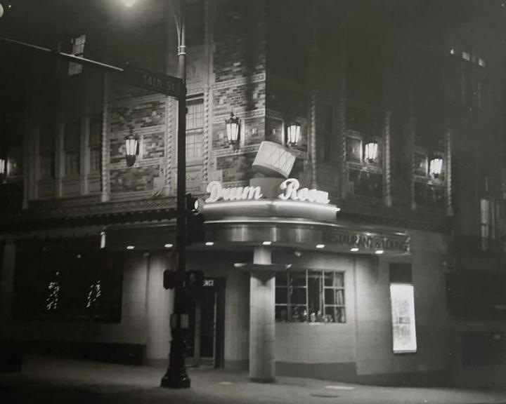 A street corner building with a neon sign for "Drum Room Restaurant & Lounge" is illuminated at night.