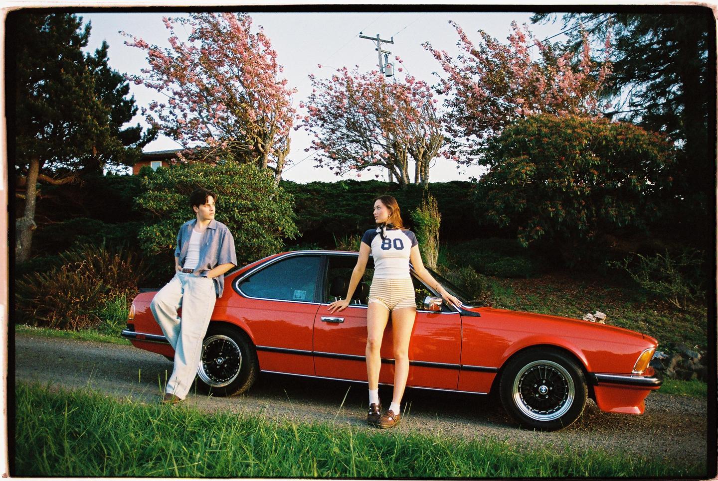 A man and a woman pose with a red car on a grassy roadside next to flowering trees.