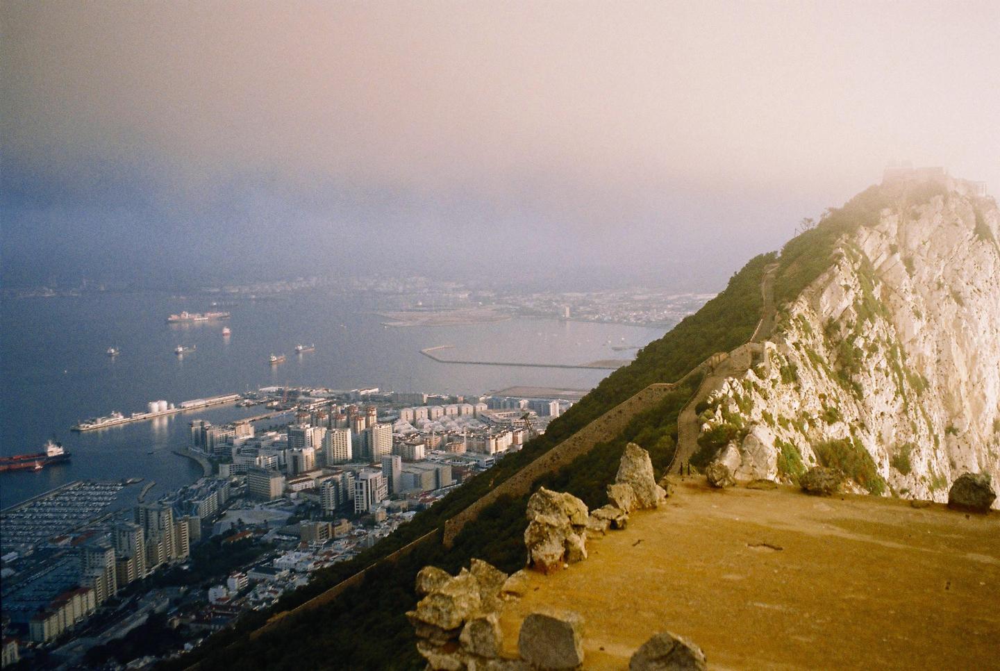 A high-angle landscape shows a city and harbor at the base of a large rock formation, with a hazy sky.