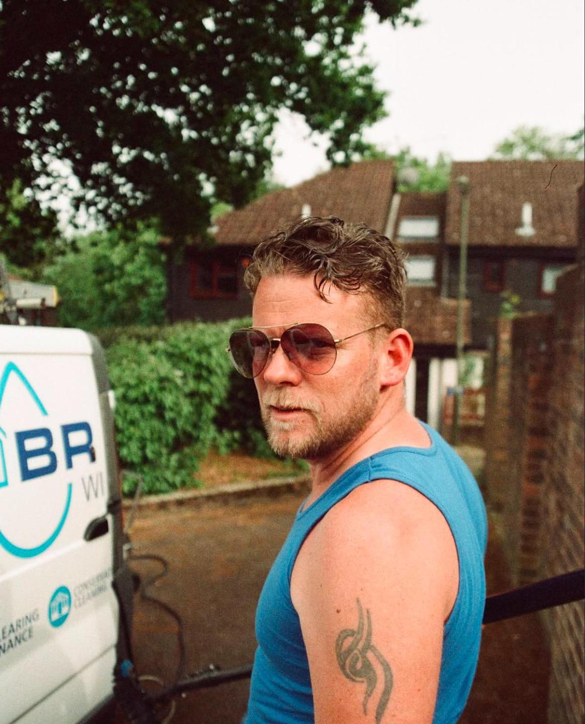 A man in a blue tank top and sunglasses looks over his shoulder with a white van and houses in the background.