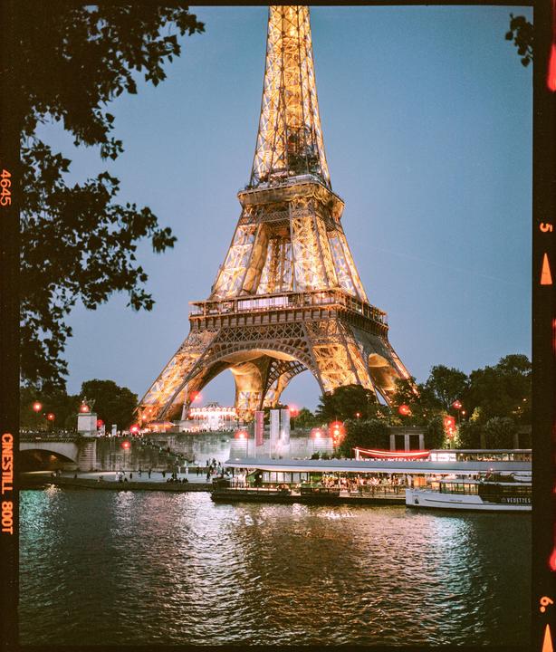 The Eiffel Tower is illuminated at night, towering over a river with boats and people on its banks.