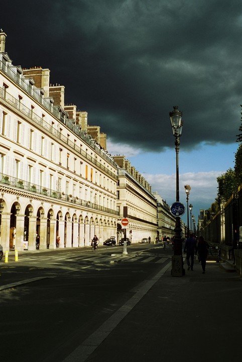 A street in Paris is lined with classical buildings featuring arched ground floors, under a dramatic sky with dark storm clouds on one side and blue sky on the other.