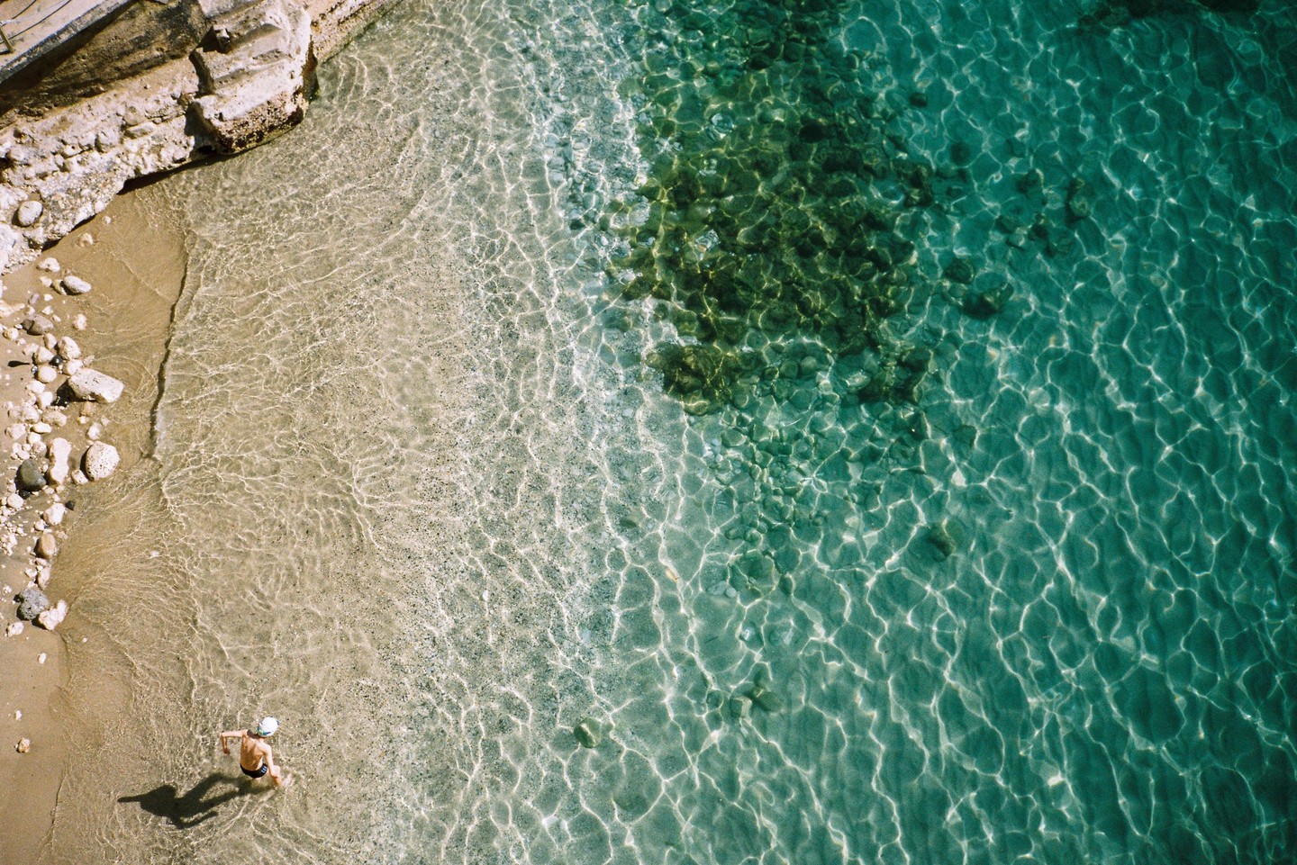 A person stands in shallow clear water at the edge of a sandy beach next to a rocky outcrop.