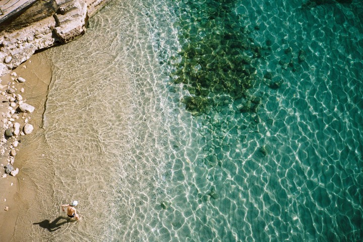 A person stands in shallow clear water at the edge of a sandy beach next to a rocky outcrop.
