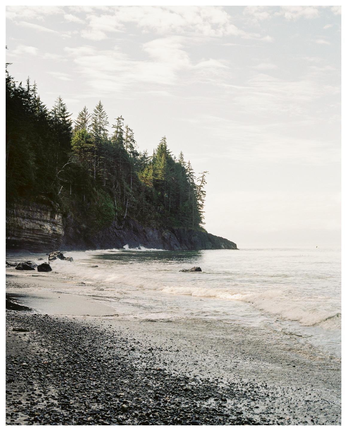 Waves crash on a rocky beach next to a cliff covered in pine trees.