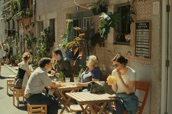 People sit at wooden tables outside a cafe on a sunny street lined with potted plants and vines.