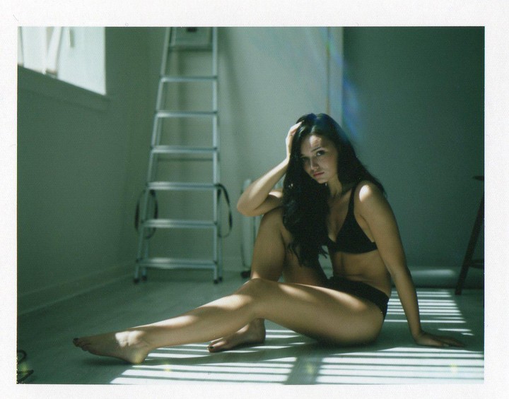 A woman in a black bra and underwear sits on a floor with striped shadows from a window.