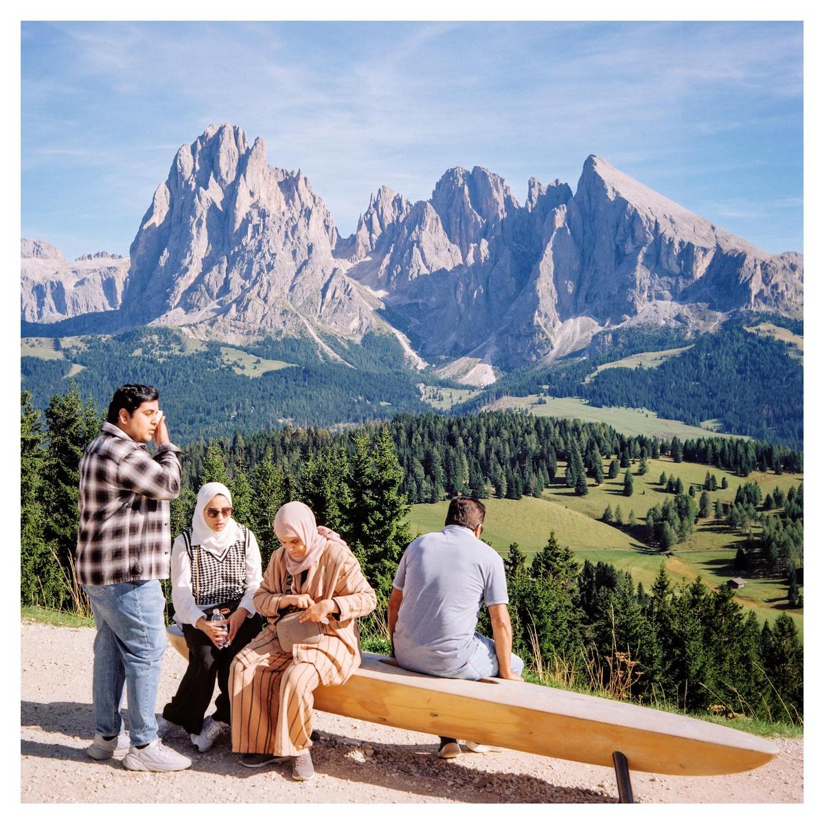 Four people are resting on a wooden bench overlooking a vast mountain landscape with forests and meadows.