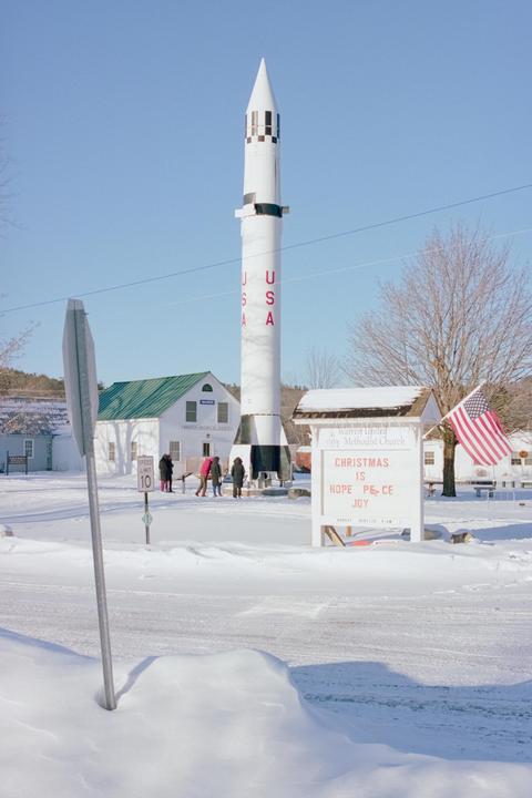 A large white rocket stands in a snow-covered village with a church sign and an American flag.