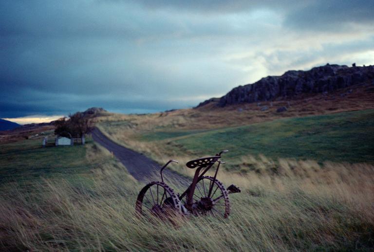 A rusty bicycle sits in tall grass next to a dirt road leading to a small building with a mountain in the background under a cloudy sky.
