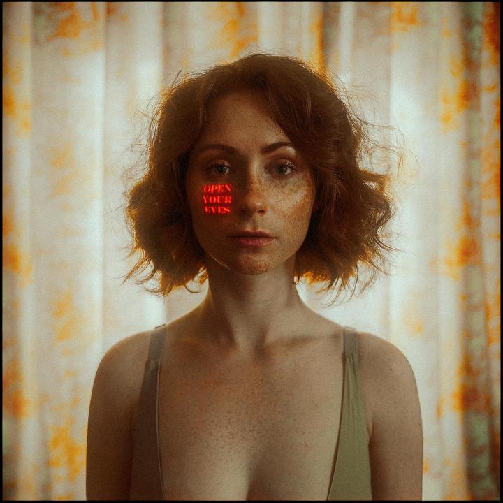 A woman with freckles and red hair looks at the viewer, with the words "OPEN YOUR EYES" projected in red light on her cheek.