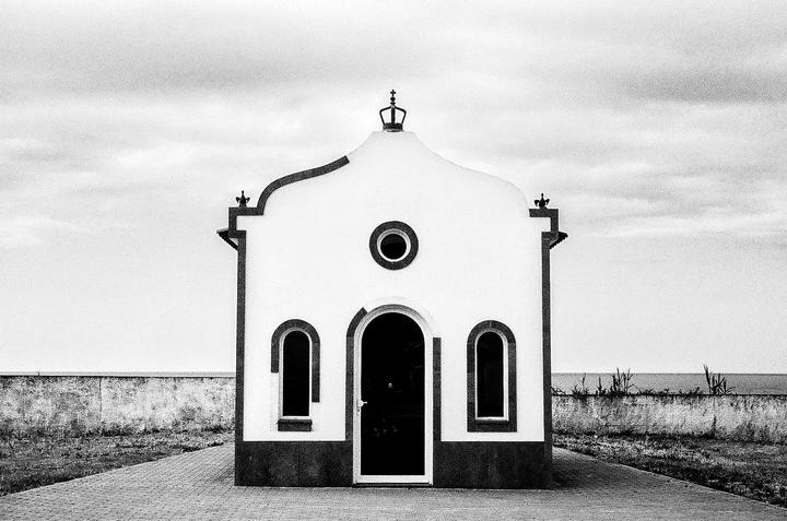 A small chapel with a crown on its roof stands on a paved path next to a low wall and the ocean under a cloudy sky.
