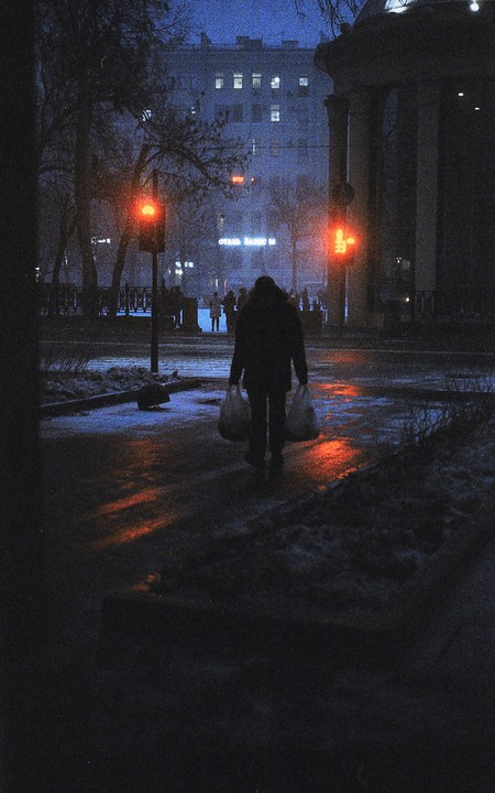 A person walks away from the camera carrying two bags on a snowy street at night, with red traffic lights illuminating the wet ground.