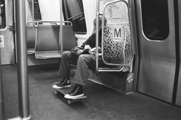 A person sits on a subway train with their feet on a skateboard.