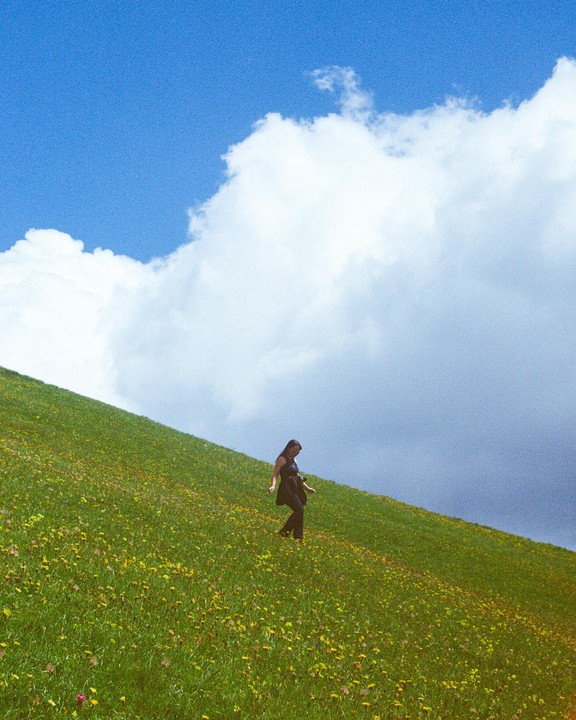 A woman walks down a grassy hill covered in yellow flowers under a bright blue sky with large white clouds.