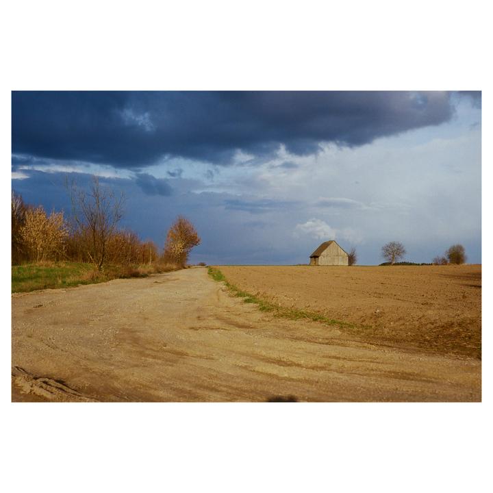 A dirt road leads through a field towards a small wooden barn under a dramatic sky.