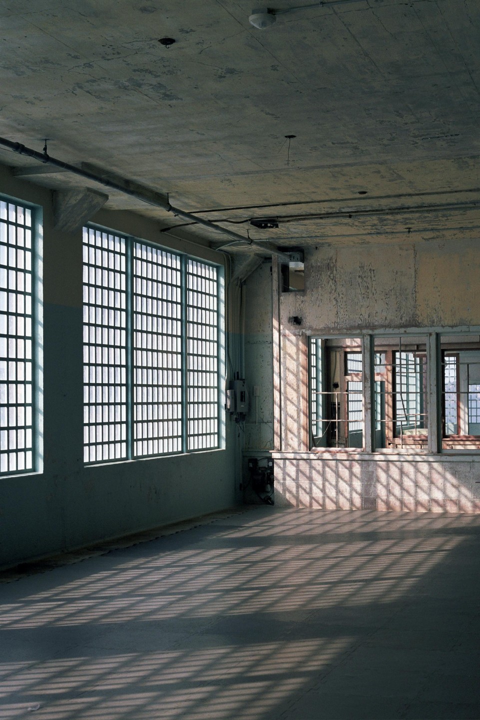 Sunlight streams through barred windows, casting grid shadows on the floor and wall of an empty room.