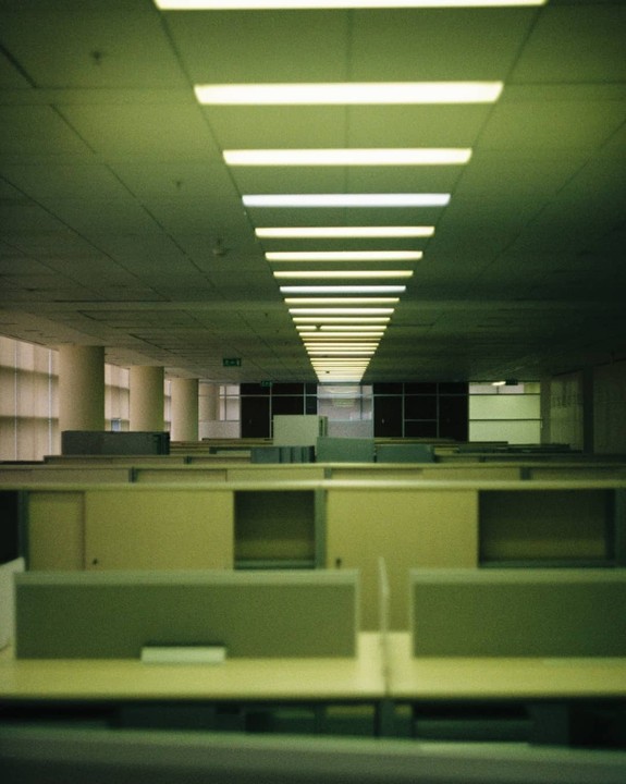 A long row of fluorescent lights illuminates an empty office space with cubicles.