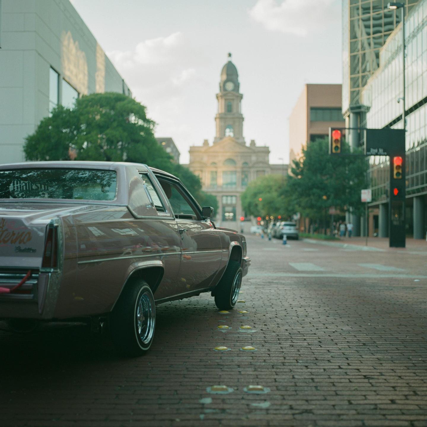 A pink lowrider car is parked on a brick street with a clock tower building in the background.