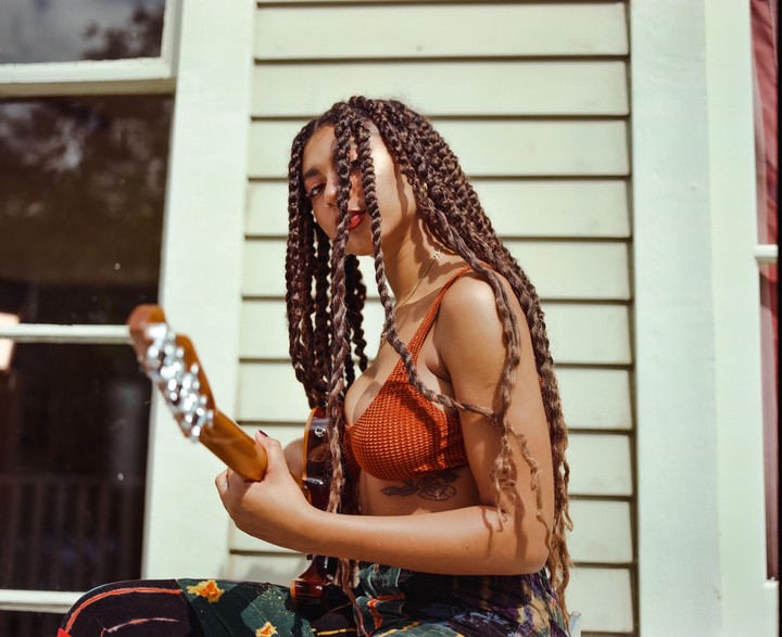 A woman with long braids holds a guitar while looking at the camera, with a light-colored wooden wall and window in the background.