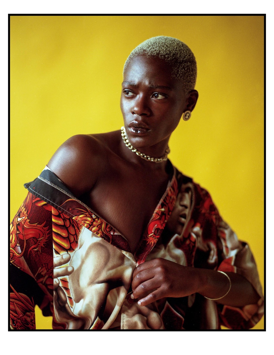 A woman with short, light-colored hair and a gold chain around her neck unbuttons a patterned shirt against a yellow background.