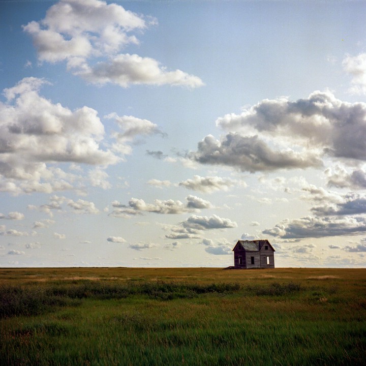 An abandoned wooden house stands alone in a vast grassy field under a cloudy sky.