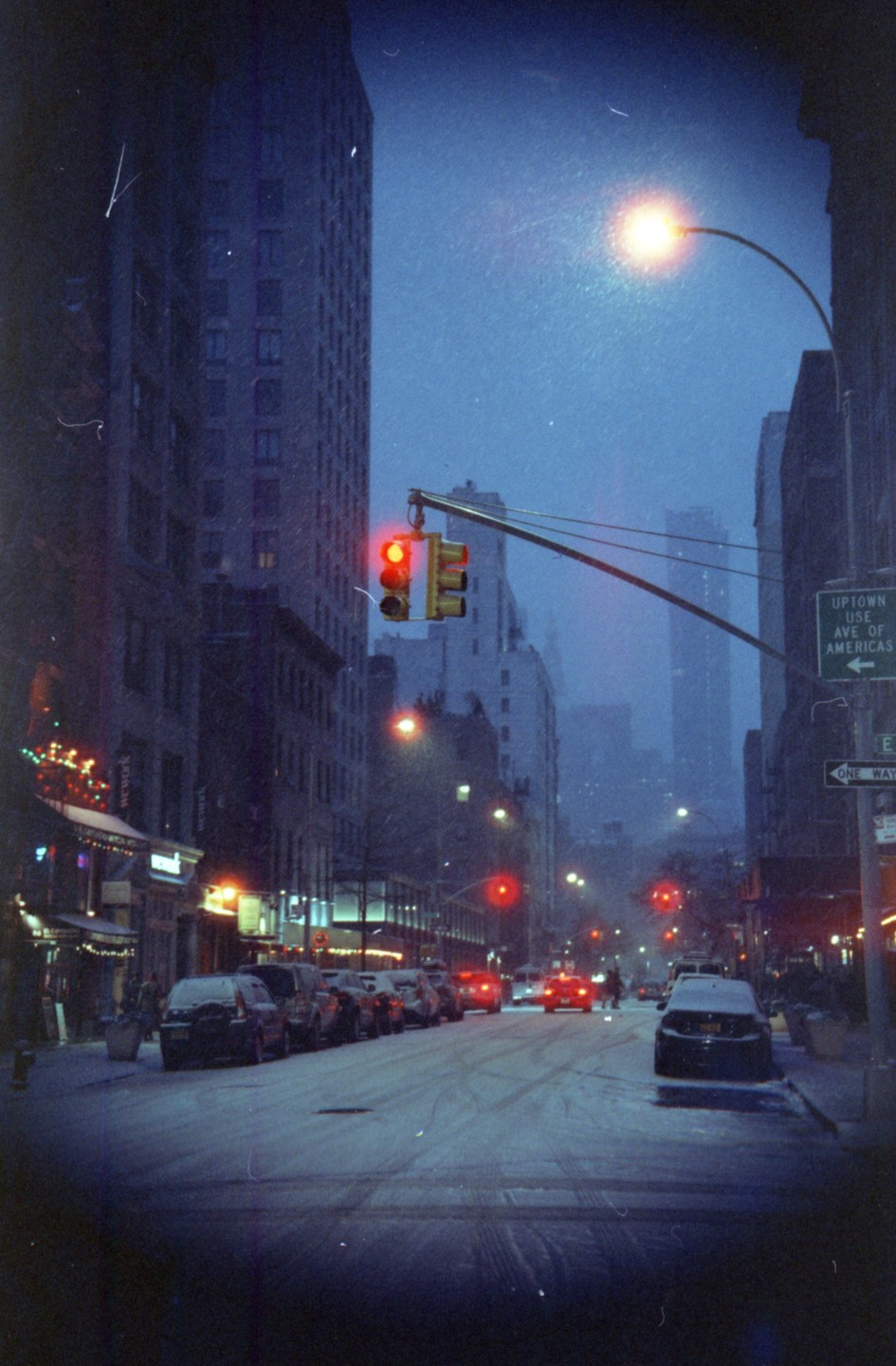 A snow-covered street in New York City is lined with parked cars and buildings under a dark, hazy sky.