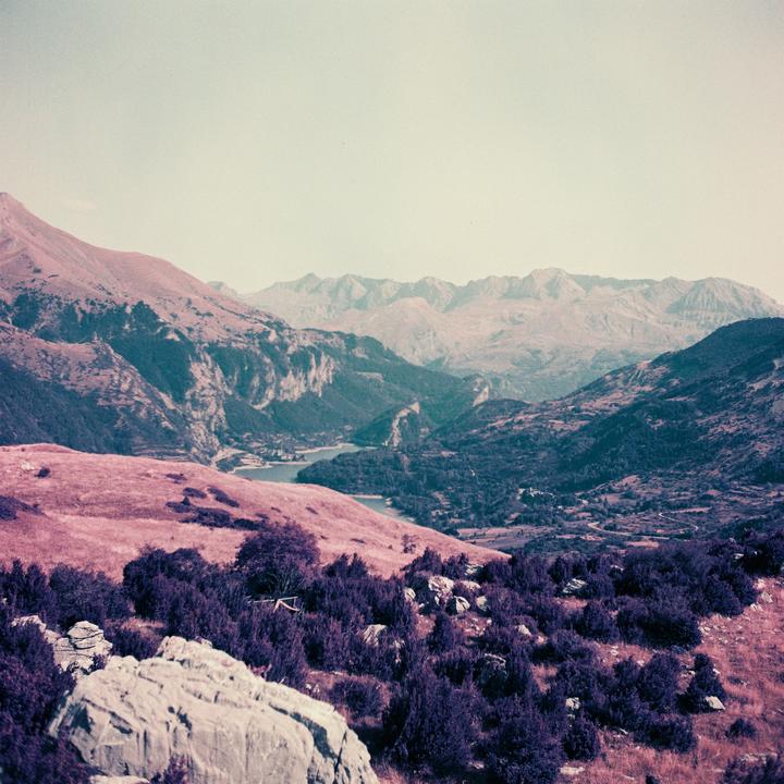 A wide landscape shows a valley with a lake surrounded by mountains and purple foliage.