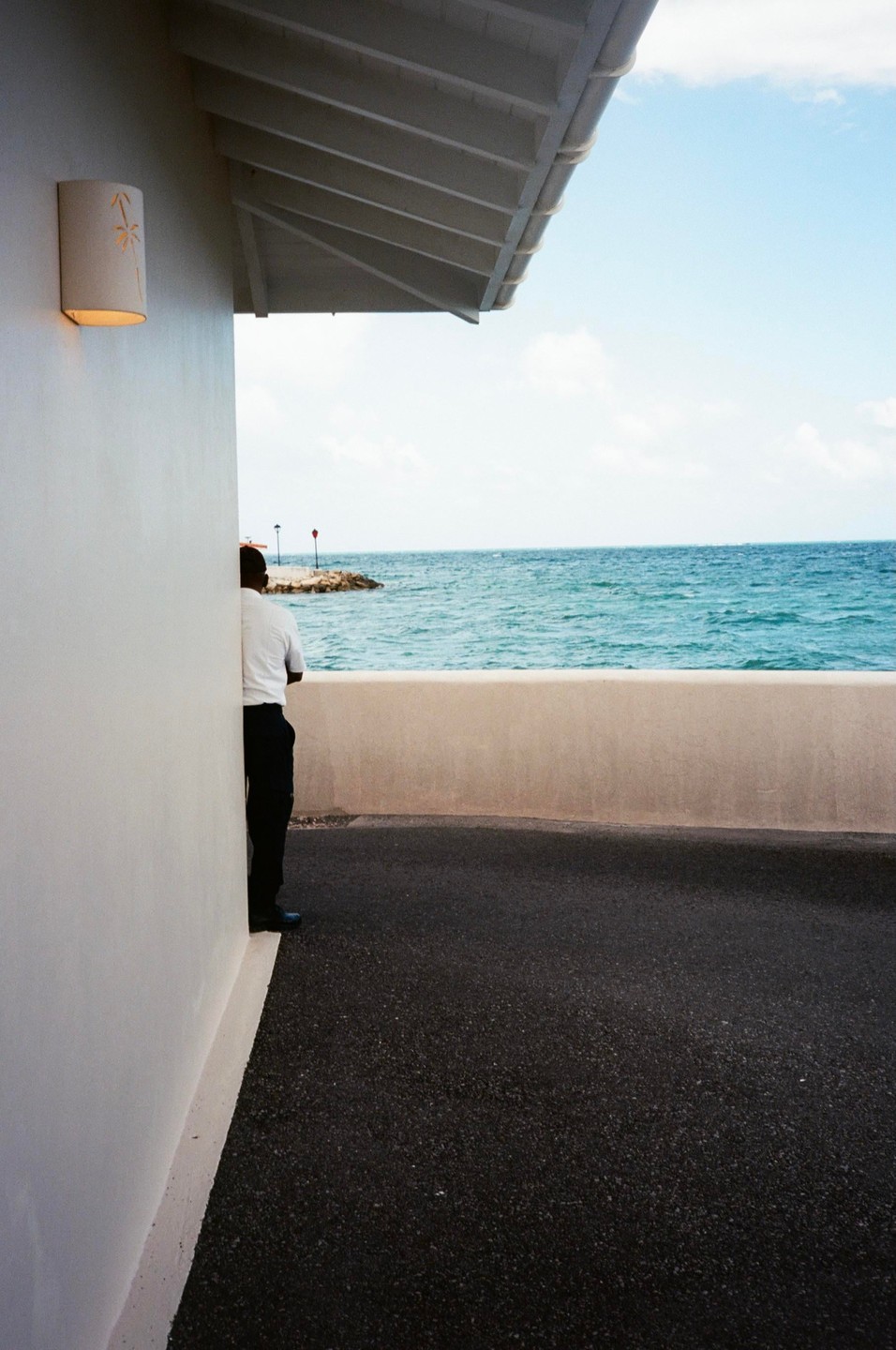 A man stands next to a white wall overlooking the ocean with a rocky pier in the distance.