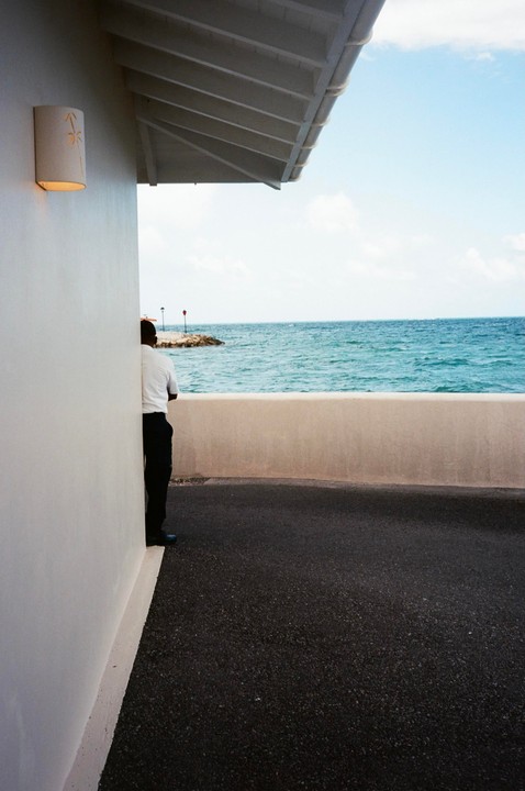 A man stands next to a white wall overlooking the ocean with a rocky pier in the distance.