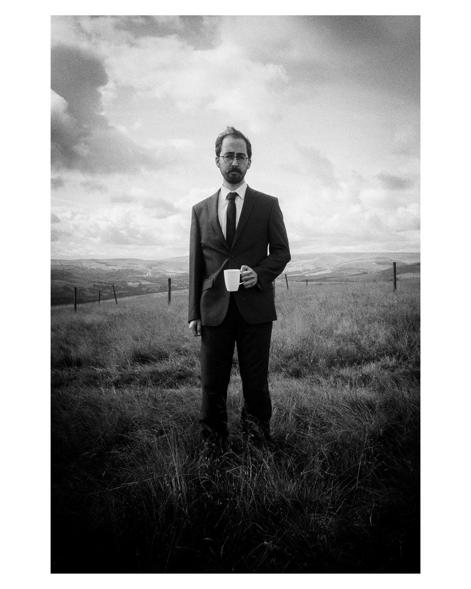 A man in a suit and tie stands in a grassy field holding a mug under a cloudy sky.