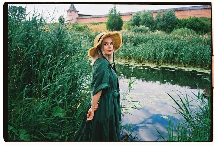 A woman in a green dress and straw hat stands by a pond with tall grass and a brick wall in the background.
