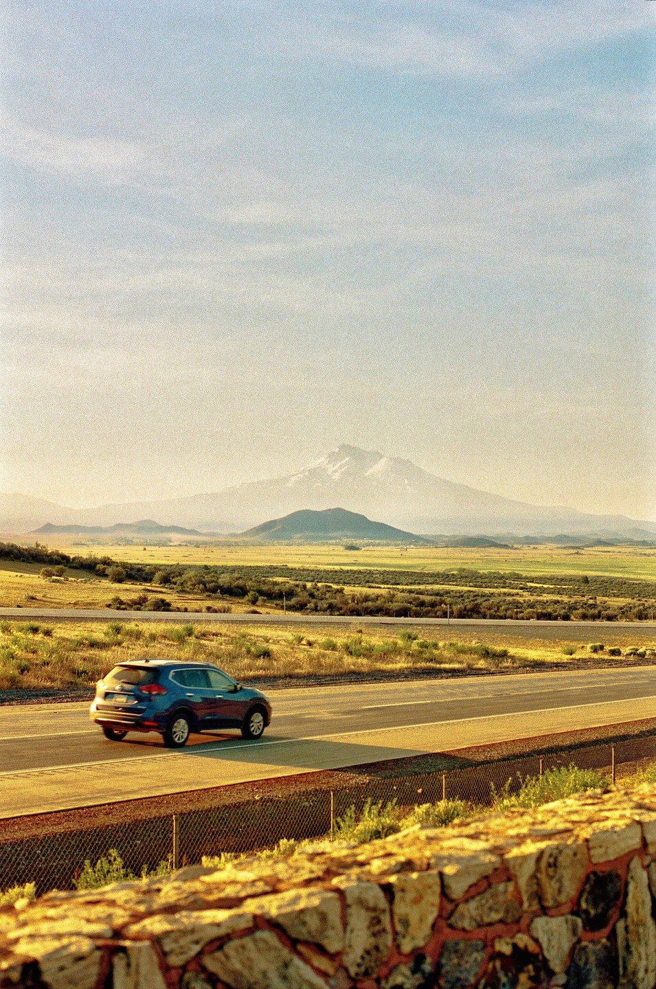A blue car drives on a highway with a snow-capped mountain in the distance under a hazy sky.