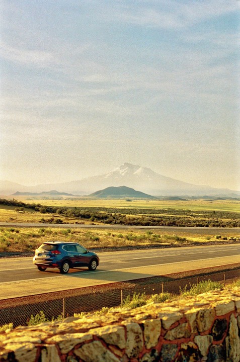 A blue car drives on a highway with a snow-capped mountain in the distance under a hazy sky.