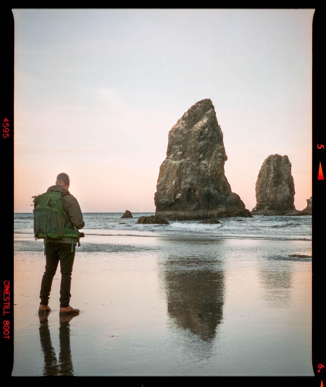 A man with a backpack stands on a wet beach looking at large sea stacks in the water.