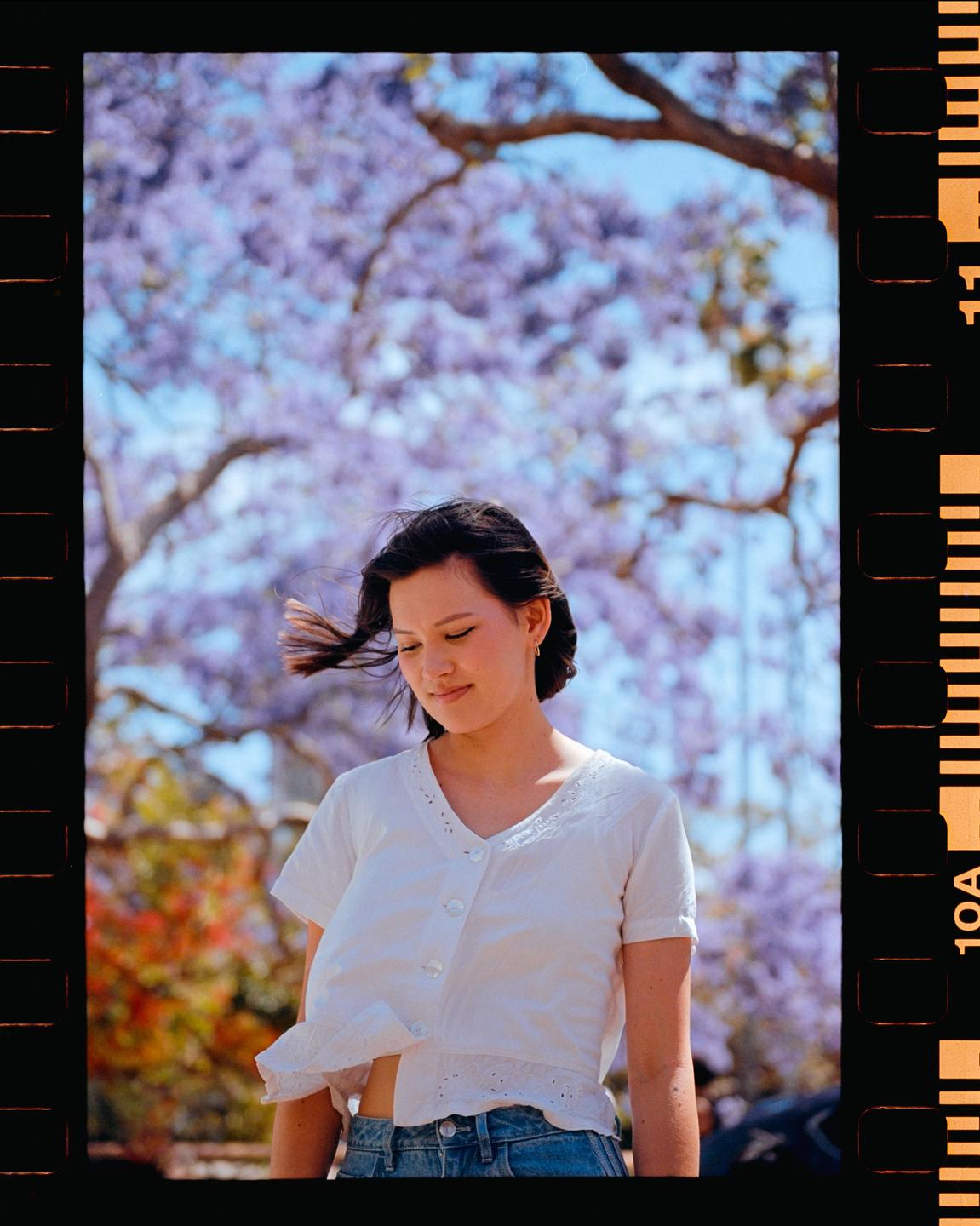 A woman stands outdoors with purple jacaranda blossoms in the background.