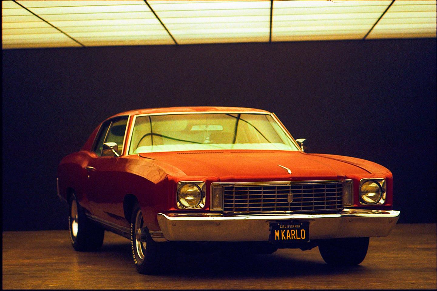 A red car with a California license plate that reads "M KARLO" is parked in a dark room with a grid of lights on the ceiling.