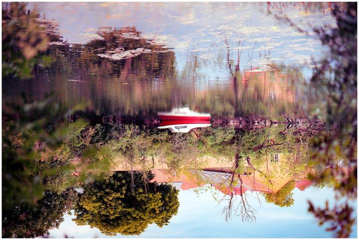 A red and white boat is docked on the shore of a body of water, with trees and buildings reflected in the water.