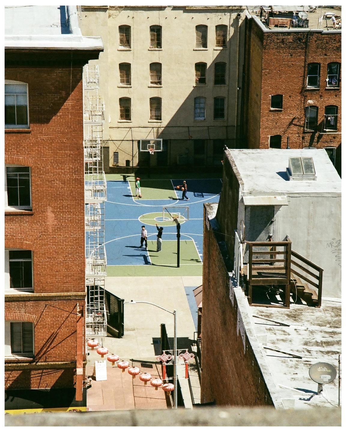 A high-angle view shows people playing basketball on a blue and green court surrounded by brick buildings in San Francisco.
