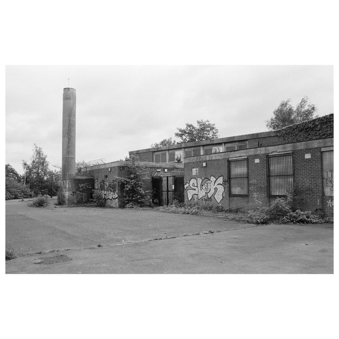 An abandoned brick building with graffiti and a tall concrete chimney stands under a cloudy sky.