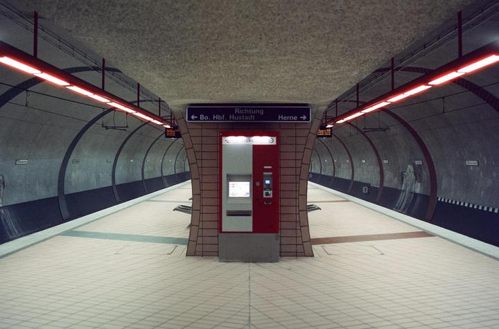 A subway platform with a ticket machine in the center is illuminated by red fluorescent lights.