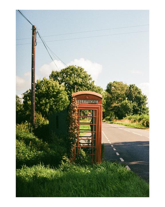 An old red telephone booth covered in vines stands next to a road with a utility pole and trees in the background.