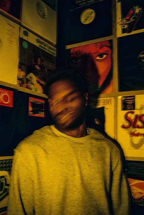 A man with a blurred face stands in front of a wall covered in record albums.