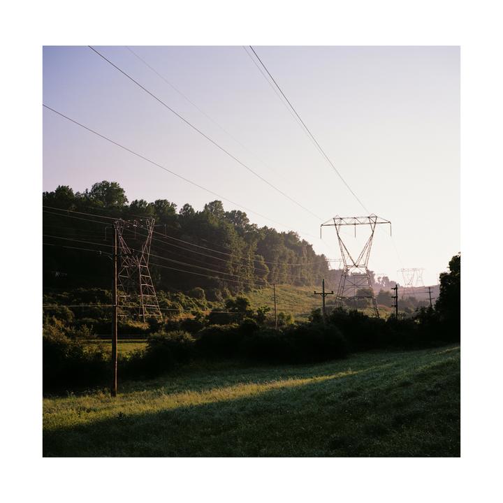 Power lines stretch across a hilly landscape with trees and a grassy field.