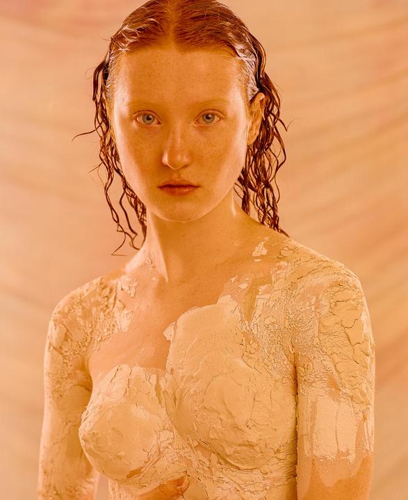 A woman with wet, curly hair and clay on her body looks directly at the viewer.
