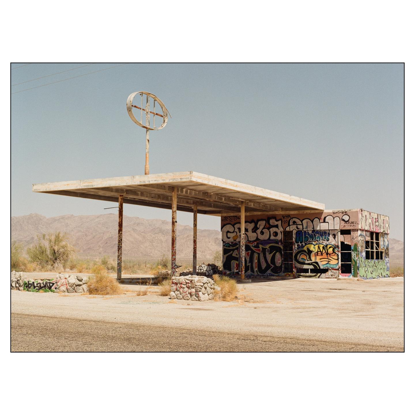 An abandoned gas station covered in graffiti stands in a desert landscape with mountains in the background.