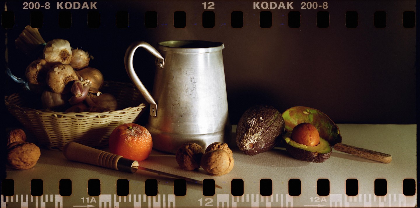 A still life arrangement features a basket of garlic and onions, a metal pitcher, an orange, walnuts, a nutcracker, and avocados, one of which is cut open.